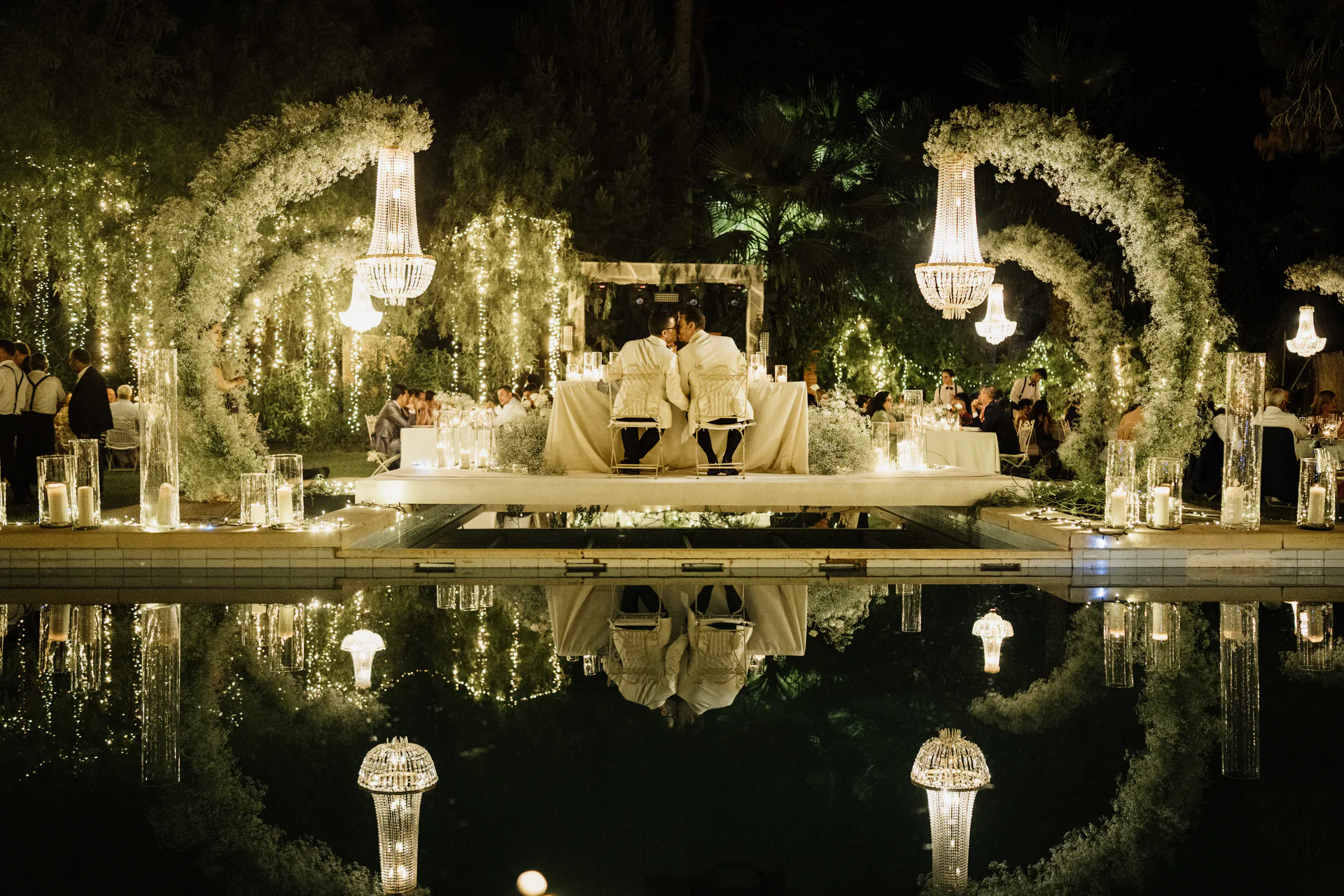 Los novios se besan en la mesa presidencial, reflejada en la piscina, en un banquete nocturno con lámparas de araña y arcos de flores